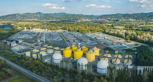 Aerial view of a large wastewater treatment and biogas production complex with yellow anaerobic digesters white domes settling tanks and solar panels in Italy.