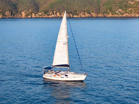 Aerial view of a white sailboat sailing on the blue waters of Lacona Bay with a rocky coastline in the background in Lacona Tuscany Italy.