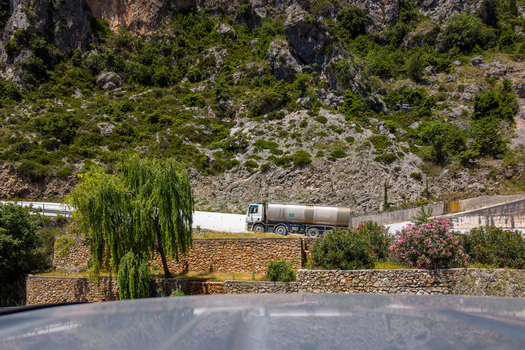 Aerial view of a white tanker truck driving along a winding mountain road past stone walls and lush green cliffs in Dhermi Vlore County Albania.