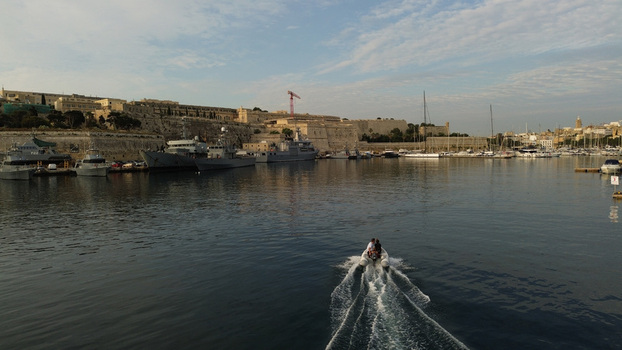 Aerial view of a small motorboat cruising through the calm waters of the Grand Harbour with historic stone fortifications and docked naval vessels in Valletta Malta. Aerial view of a small motorboat cruising through the calm waters of the Grand Harbour with historic stone fortifications and docked naval vessels in Valletta Malta.
