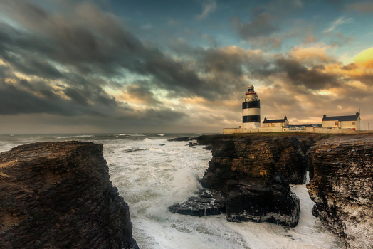 View of Hook Lighthouse standing on rocky cliffs with crashing waves under a dramatic cloudy sky at sunset in County Wexford Ireland. View of Hook Lighthouse standing on rocky cliffs with crashing waves under a dramatic cloudy sky at sunset in County Wexford Ireland.