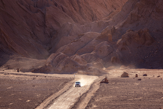 Aerial view of a white vehicle driving along a dusty road through the rugged arid terrain of the Valley of the Moon in San Pedro de Atacama Antofagasta Chile. Aerial view of a white vehicle driving along a dusty road through the rugged arid terrain of the Valley of the Moon in San Pedro de Atacama Antofagasta Chile.