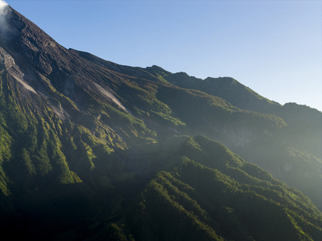 Aerial view of the rugged volcanic slopes of Mount Merapi with lush green vegetation and rocky ridges under a clear blue sky in Kecamatan Pakem Daerah Istimewa Yogya Indonesia. Aerial view of the rugged volcanic slopes of Mount Merapi with lush green vegetation and rocky ridges under a clear blue sky in Kecamatan Pakem Daerah Istimewa Yogya Indonesia.
