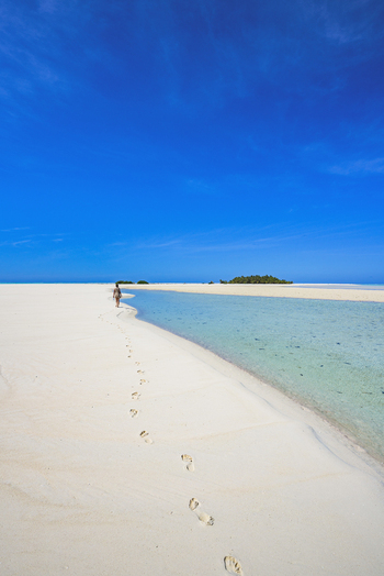 View of a person walking along the white sand beach of Aitutaki Lagoon with clear turquoise water and a bright blue sky in Aitutaki Cook Islands. View of a person walking along the white sand beach of Aitutaki Lagoon with clear turquoise water and a bright blue sky in Aitutaki Cook Islands.