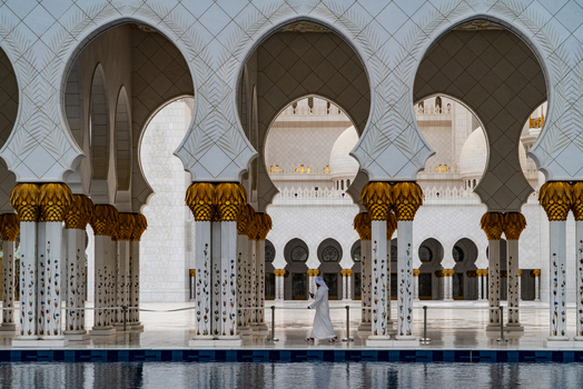 View of the white marble arches and intricate floral patterns of a grand mosque with a man walking through the courtyard in Abu Dhabi Abu Dhabi United Arab Emirates. View of the white marble arches and intricate floral patterns of a grand mosque with a man walking through the courtyard in Abu Dhabi Abu Dhabi United Arab Emirates.