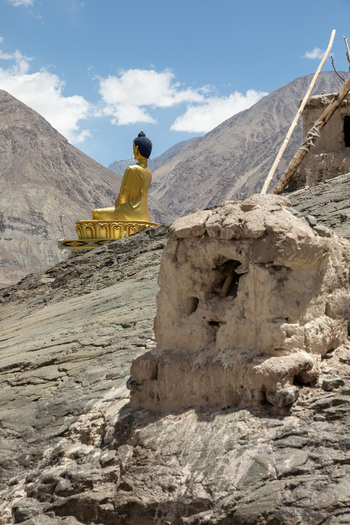 View of the golden Buddha statue at Charasa Gonpa set against a backdrop of rugged mountains and ancient stone ruins in the arid landscape of Charasa Ladakh India.