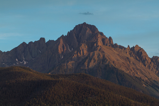 Aerial view of Mount Sneffels illuminated by golden hour light rising above the dense evergreen forests of the San Juan Mountain Range in Ouray Colorado United States. Aerial view of Mount Sneffels illuminated by golden hour light rising above the dense evergreen forests of the San Juan Mountain Range in Ouray Colorado United States.