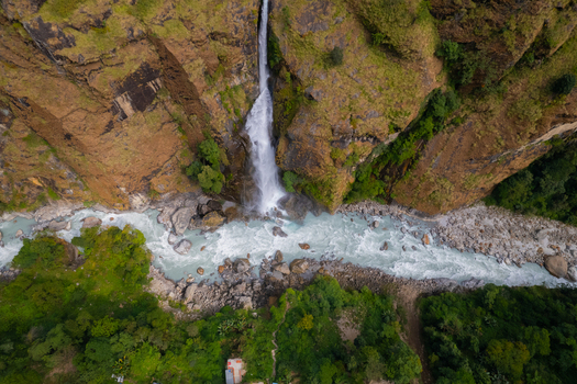 Aerial view of a waterfall cascading down a steep rocky cliff into a turquoise river surrounded by lush green forest and rocky banks in Nepal.