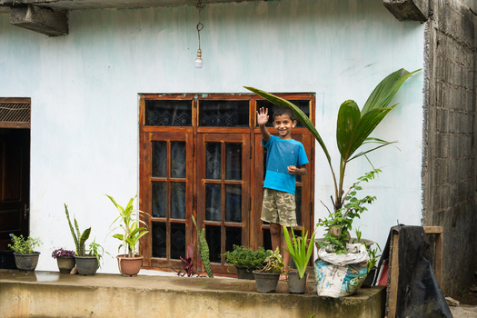 Sri Lanka 20 February 2026 View of a young boy waving his hand while standing on a porch decorated with potted plants in front of a modest house.