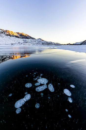 View of a frozen lake with trapped air bubbles in the foreground and snow covered mountains under a clear sky at sunset Madesimo Lombardy Italy. View of a frozen lake with trapped air bubbles in the foreground and snow covered mountains under a clear sky at sunset Madesimo Lombardy Italy.