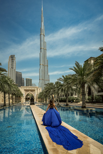 Dubai United Arab Emirates 24 April 2023 View of the Burj Khalifa with a woman in a long blue dress standing on a stone path by a reflecting pool under a clear blue sky. Dubai United Arab Emirates 24 April 2023 View of the Burj Khalifa with a woman in a long blue dress standing on a stone path by a reflecting pool under a clear blue sky.