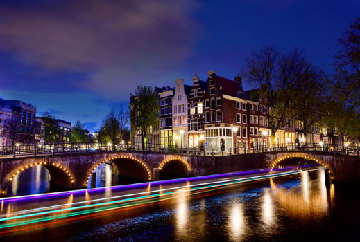 View of historic canal houses illuminated at night along a bridge with light trails from a passing boat in the Netherlands. View of historic canal houses illuminated at night along a bridge with light trails from a passing boat in the Netherlands.