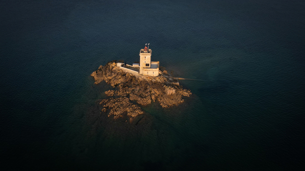Aerial view of the stark white lighthouse stands defiant on Ile Noire amidst the dark swirling sea contrasting light and shadow Plouezoc h Finistere France.