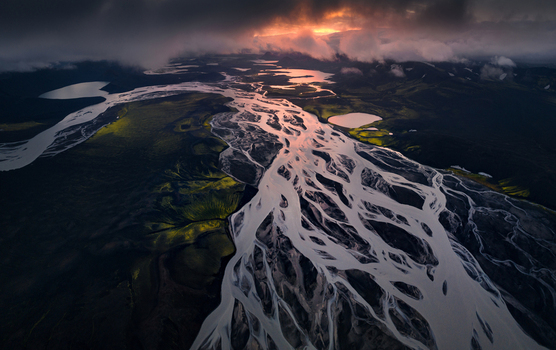 Aerial view of a braided glacial river system flowing through a dark volcanic landscape with mossy hills under a dramatic sunset sky in Iceland.