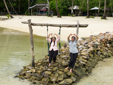 Aerial view of two women playfully pose on a swing set amidst the rocky shore framed by swaying palm trees and tranquil waters at Bobby Beach Karimunjawa Central Java Indonesia.