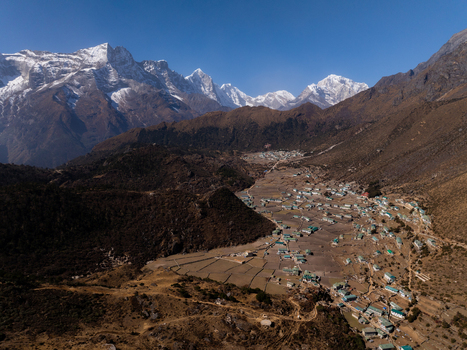 Aerial view of a remote Himalayan village nestled in a valley with terraced fields and small houses surrounded by towering snow capped mountain peaks in Nepal.