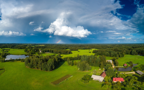 Aerial view of lush green fields dense forests small ponds and traditional farmhouses under a dramatic sky with a faint rainbow in Latvia. Aerial view of lush green fields dense forests small ponds and traditional farmhouses under a dramatic sky with a faint rainbow in Latvia.