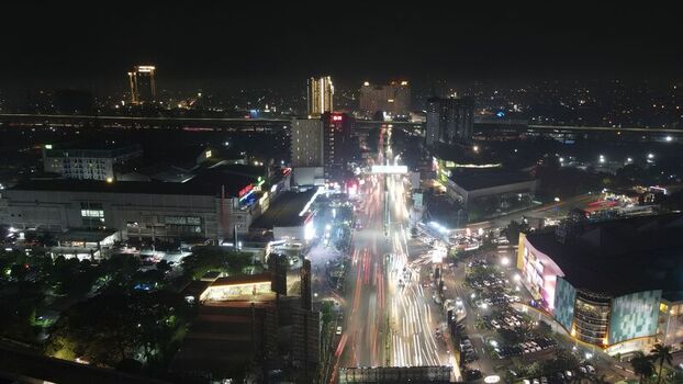 Aerial view of illuminated city streets with long exposure light trails from traffic surrounded by urban buildings and parking areas in Bekasi Barat West Java Indonesia. Aerial view of illuminated city streets with long exposure light trails from traffic surrounded by urban buildings and parking areas in Bekasi Barat West Java Indonesia.