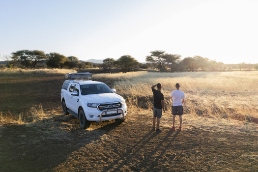 Aerial view of two men standing near a white vehicle on dry land with sparse trees under the bright sun Windhoek Namibia. Aerial view of two men standing near a white vehicle on dry land with sparse trees under the bright sun Windhoek Namibia.