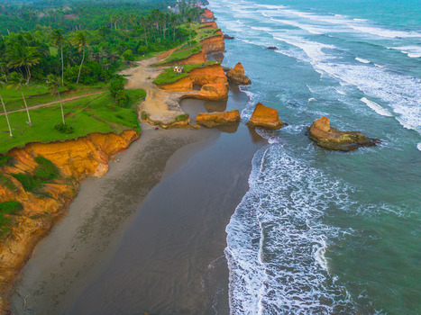 Aerial view of dramatic orange cliffs and sandy beach meeting the turquoise ocean waves with lush palm trees along the coastline in Bali Bali Indonesia. Aerial view of dramatic orange cliffs and sandy beach meeting the turquoise ocean waves with lush palm trees along the coastline in Bali Bali Indonesia.