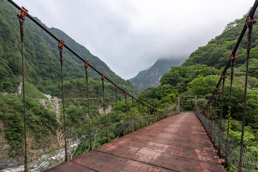 View of the suspension bridge at Taroko National Park with metal decking spanning a gorge surrounded by lush green mountains under a cloudy sky in Xiulin Township Hualien County Taiwan.
