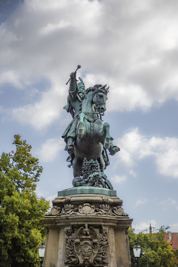 View of the King John III Sobieski Monument a bronze equestrian statue on an ornate stone pedestal surrounded by lush green trees under a cloudy sky in Gdansk Pomeranian Voivodeship Poland. View of the King John III Sobieski Monument a bronze equestrian statue on an ornate stone pedestal surrounded by lush green trees under a cloudy sky in Gdansk Pomeranian Voivodeship Poland.