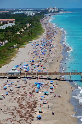 Aerial view of Juno Beach Pier and a crowded sandy coastline with turquoise ocean water colorful umbrellas and people enjoying the sunny day in Juno Beach Florida United States. Aerial view of Juno Beach Pier and a crowded sandy coastline with turquoise ocean water colorful umbrellas and people enjoying the sunny day in Juno Beach Florida United States.