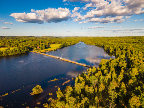 Aerial view of a long bridge crossing a calm lake surrounded by dense green forests under a blue sky with scattered clouds in Gallared Halland County Sweden. Aerial view of a long bridge crossing a calm lake surrounded by dense green forests under a blue sky with scattered clouds in Gallared Halland County Sweden.