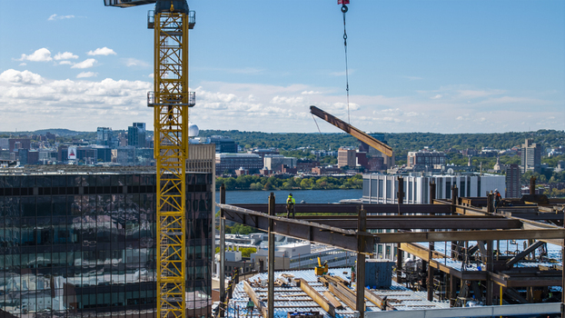 Aerial view of Kendall Square construction site with a yellow tower crane lifting a steel beam workers on the frame and city skyline in Cambridge Massachusetts United States. Aerial view of Kendall Square construction site with a yellow tower crane lifting a steel beam workers on the frame and city skyline in Cambridge Massachusetts United States.