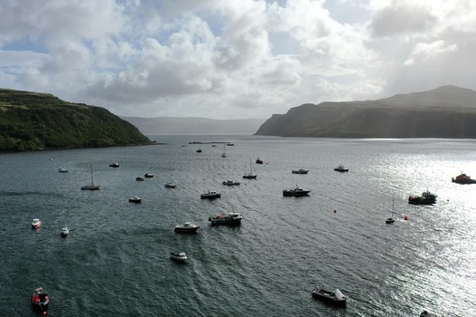 Aerial view of Portree Harbour with numerous small fishing boats and yachts anchored in the bay surrounded by green hills under a cloudy sky in Portree Scotland United Kingdom.