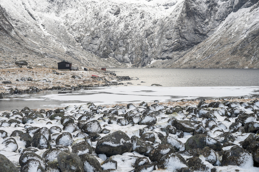 View of a snowy landscape with a frozen lake dark rocks in the foreground and a small cabin nestled at the base of rugged mountains in Leknes Lofoten Norway.