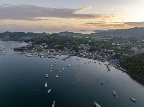 Aerial view of Labuan Bajo harbor with boats anchored in the bay surrounded by coastal town buildings green hills and a calm sea under a sunset sky in Komodo East Nusa Tenggara Indonesia. Aerial view of Labuan Bajo harbor with boats anchored in the bay surrounded by coastal town buildings green hills and a calm sea under a sunset sky in Komodo East Nusa Tenggara Indonesia.
