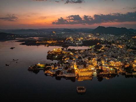 Aerial view of the illuminated City Palace and historic buildings along the shores of Lake Pichola at twilight in Udaipur Rajasthan India.