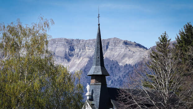 Aerial view of the Chapelle Notre Dame de Riantmont spire with rocky mountain peaks and green trees under a clear blue sky in Vesancy Auvergne Rhone Alpes France. Aerial view of the Chapelle Notre Dame de Riantmont spire with rocky mountain peaks and green trees under a clear blue sky in Vesancy Auvergne Rhone Alpes France.