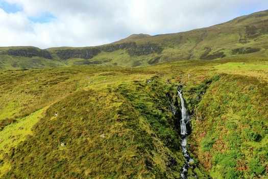 Aerial view of Bride s Veil Falls cascading down a lush green hillside with rolling grassy terrain under a cloudy sky Portree Scotland United Kingdom. Aerial view of Bride s Veil Falls cascading down a lush green hillside with rolling grassy terrain under a cloudy sky Portree Scotland United Kingdom.