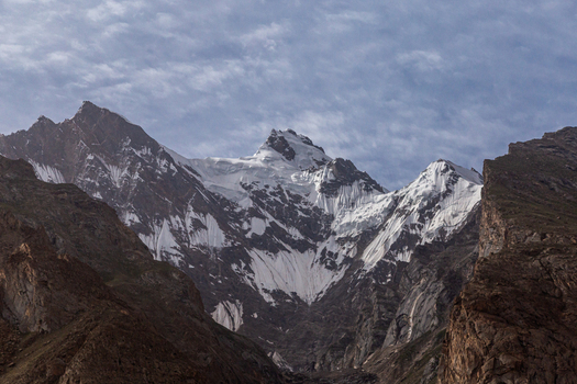 View of the snow capped peaks of Baltoro Glacier rising above rugged rocky slopes under a cloudy sky in Baltoro Gilgit Baltistan Pakistan.