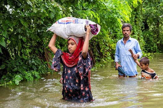 Banshkhali Upazila Chittagong Division Bangladesh 10 July 2026 View of a young girl carrying a sack on her head while wading through floodwaters with a man and a boy in the background. Banshkhali Upazila Chittagong Division Bangladesh 10 July 2026 View of a young girl carrying a sack on her head while wading through floodwaters with a man and a boy in the background.