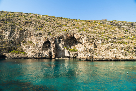 View of the turquoise waters of Xlendi Bay framed by rugged limestone cliffs under a clear blue sky in Zebbug Gozo Region Malta.