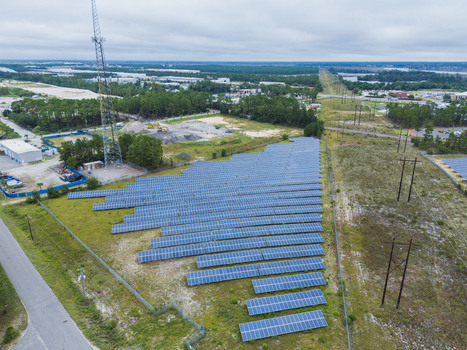 Aerial view of a solar farm with rows of photovoltaic panels installed on a grassy field featuring a communication tower and surrounding trees in Wilmington North Carolina United States. Aerial view of a solar farm with rows of photovoltaic panels installed on a grassy field featuring a communication tower and surrounding trees in Wilmington North Carolina United States.