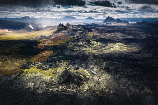 Aerial view of the rugged volcanic landscape featuring dark lava fields mossy craters and geothermal steam under a dramatic sky with light rays Iceland.