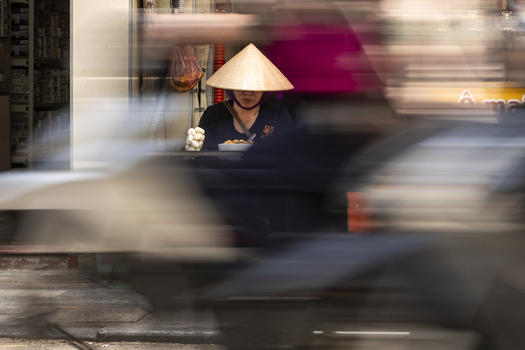 View of a woman wearing a traditional Vietnamese conical hat eating a meal at a street stall framed by the motion blur of passing motorbikes in Hanoi Ha Noi Vietnam.