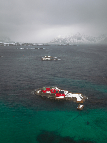 Aerial view of red buildings on a rocky island contrast against the deep blue sea and snow dusted landscape Svolv&aelig;r Nordland Norway.