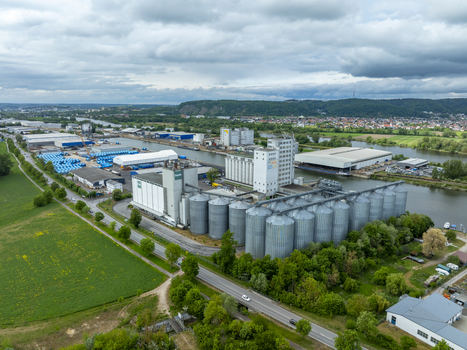 Aerial view of a large industrial grain silo complex and warehouse facility situated along a river canal with green fields and a cloudy sky.