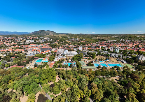 Aerial view of outdoor pools and a thermal bath complex beside a leafy park with red roofed city center and surrounding hills in Eger Hungary on a clear summer day. Aerial view of outdoor pools and a thermal bath complex beside a leafy park with red roofed city center and surrounding hills in Eger Hungary on a clear summer day.