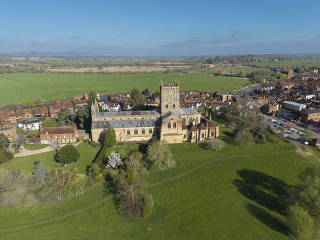 Aerial view of Tewkesbury Abbey surrounded by lush green fields historic town rooftops and trees under a clear blue sky in Tewkesbury England United Kingdom.