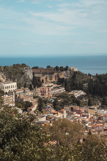 View of sun drenched buildings nestled amongst lush greenery cascading toward the azure sea under a hazy sky creating a picturesque scene Taormina Sicily Italy.