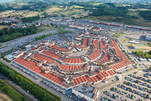 Aerial view of Designer Outlet Serravalle featuring red tiled roofs and expansive parking lots surrounded by green hills in Serravalle Scrivia Piemonte Italy. Aerial view of Designer Outlet Serravalle featuring red tiled roofs and expansive parking lots surrounded by green hills in Serravalle Scrivia Piemonte Italy.