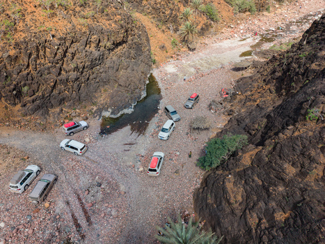 Aerial view of cars parked near a stream winding through rugged arid terrain with dramatic cliffs casting long shadows Dirhur canyon Socotra Yemen.