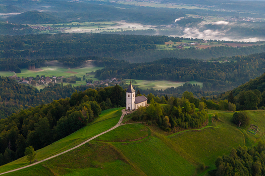 Aerial view of Church of Saints Primus and Felician situated on a grassy hill surrounded by dense forests and misty valleys in the early morning light Jamnik Kranj Slovenia.