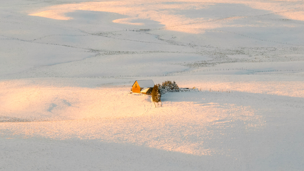 Aerial view of a solitary typical buron cabin glows warmly amidst a vast undulating expanse of pristine snow kissed by the golden light of the setting sun Nasbinals Occitanie France.
